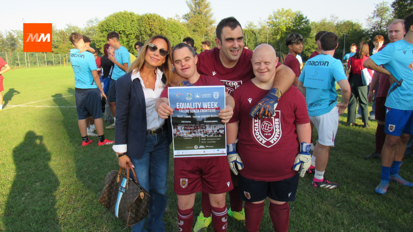 Rappresentante di MM Operations e atleti della Regia For Special di AC Reggiana sorridono insieme sul campo da calcio durante l'Equality Week, tenendo una locandina dell'evento "Calcio Senza Frontiere".