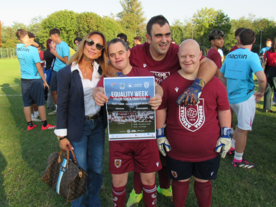 Rappresentante di MM Operations e atleti della Regia For Special di AC Reggiana sorridono insieme sul campo da calcio durante l'Equality Week, tenendo una locandina dell'evento "Calcio Senza Frontiere".
