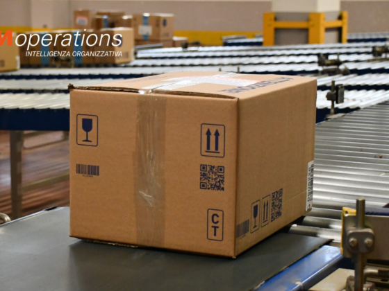 Collo in cartone con barcode, QR code e simboli di movimentazione su un nastro trasportatore a rulli in un magazzino MM Operations: identificazione del dato alla fonte per kpi logistici